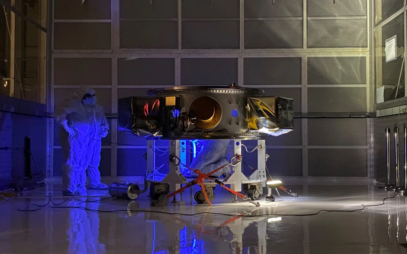 The integrated flight system on its handling dolly under blue inspection lighting, with a technician in cleanroom coveralls alongside.